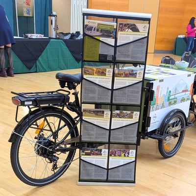 The TREC-Cycle (an electric cargo tricycle with PSU-themed artwork) parked in the PSU Smith Ballroom next to a rack of informational one-pagers.