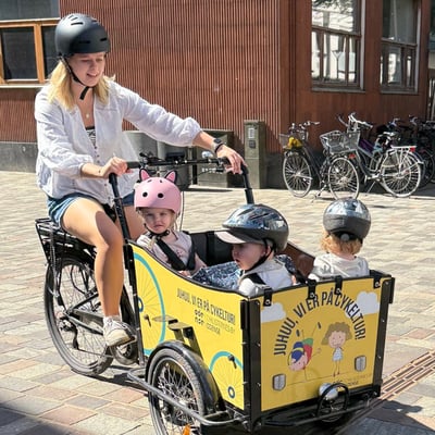 An adult rides a cargo bike with three children in the cargo compartment. All are wearing helmets.